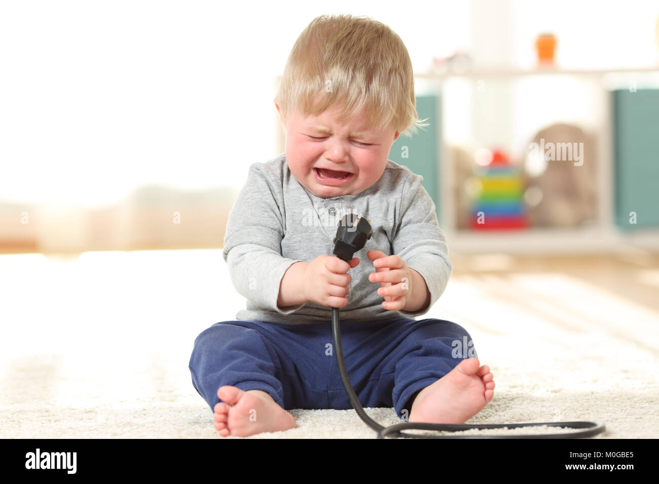 Vista anteriore verticale di un bambino aby in pericolo il pianto di trattenimento di un una spina elettrica seduta sul pavimento a casa Foto Stock