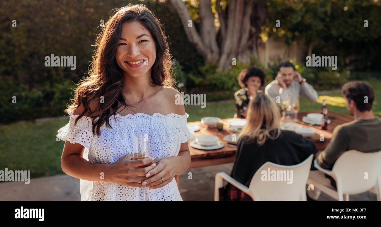Bella donna asiatica in piedi all'aperto con gli amici seduti in sfondo avente il cibo. Donna con un drink al party all'aperto. Foto Stock