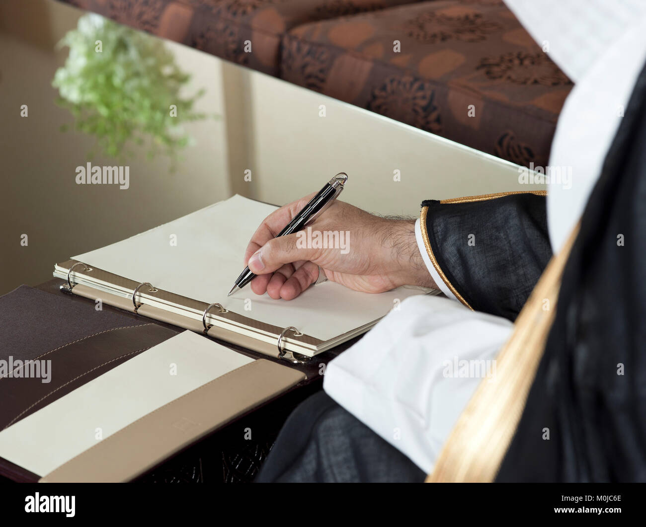 Saudi Arabian man mano che scrive su un notebook in un lussuoso ambiente domestico, indossando Arabia Thob, Ghutra e Bisht nero Foto Stock
