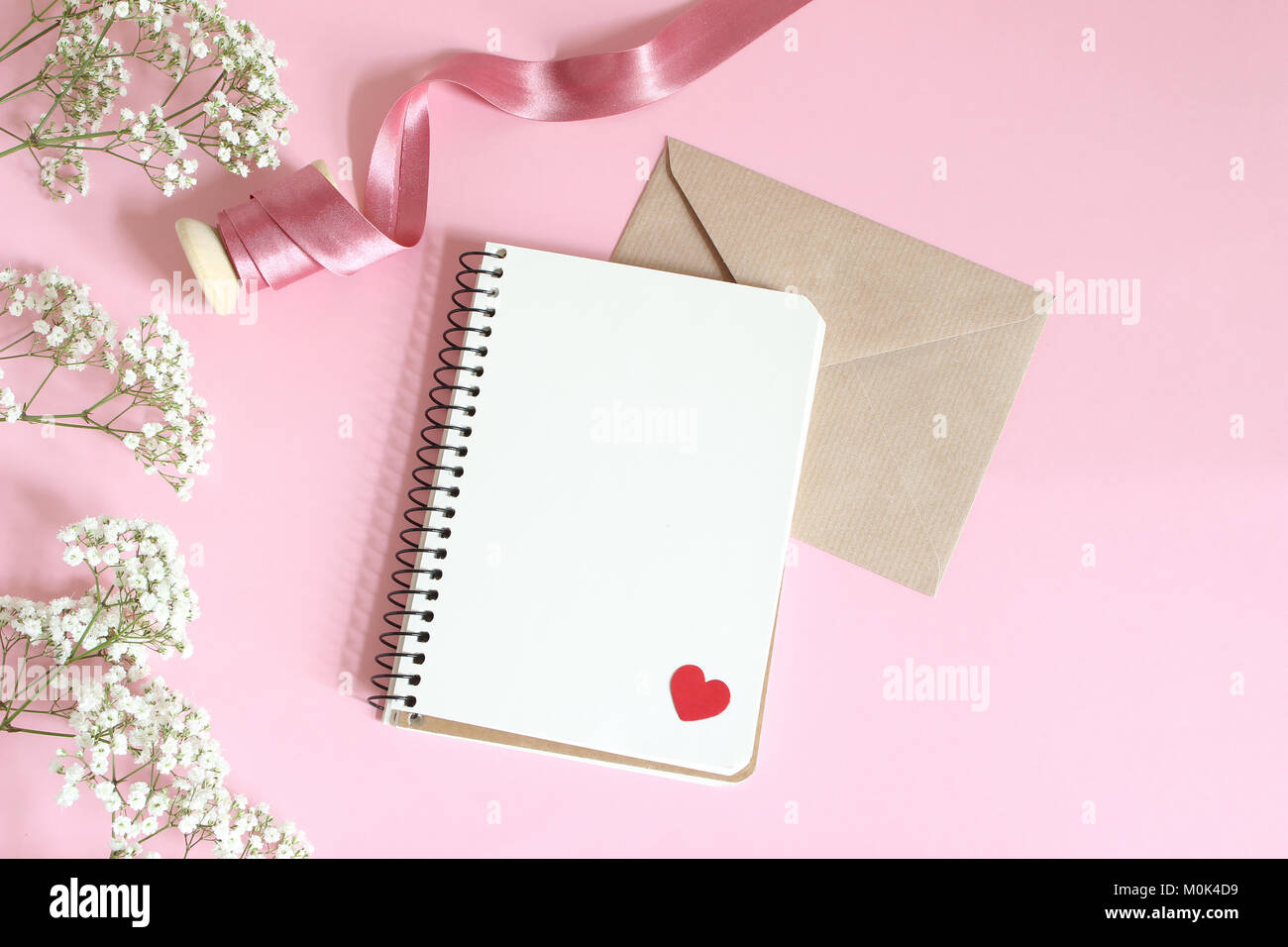 Matrimoni, compleanni o Valentines stationery mockup. White baby soffio Gypsophila fiori, nastro di raso, craft busta di carta vuoto e notebook, sfondo rosa. Stile femminile stock photo. Lay piatto. Foto Stock