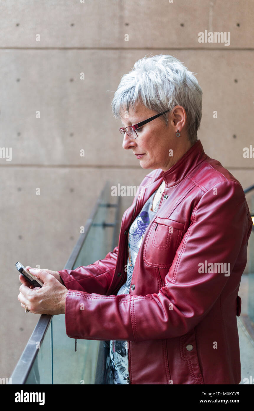 Una donna matura in un rosso giacca di pelle si trova di fronte a una ringhiera di protezione utilizzando un telefono intelligente; Vancouver, British Columbia, Canada Foto Stock