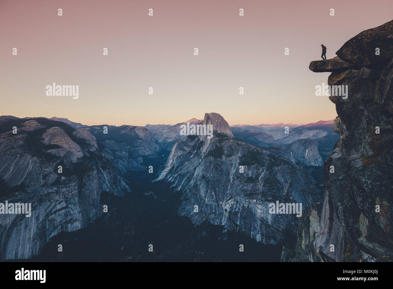 Un intrepido escursionista è in piedi su una roccia a strapiombo guardando verso il famoso Half Dome nel Parco Nazionale di Yosemite nel crepuscolo al tramonto, CALIFORNIA, STATI UNITI D'AMERICA Foto Stock