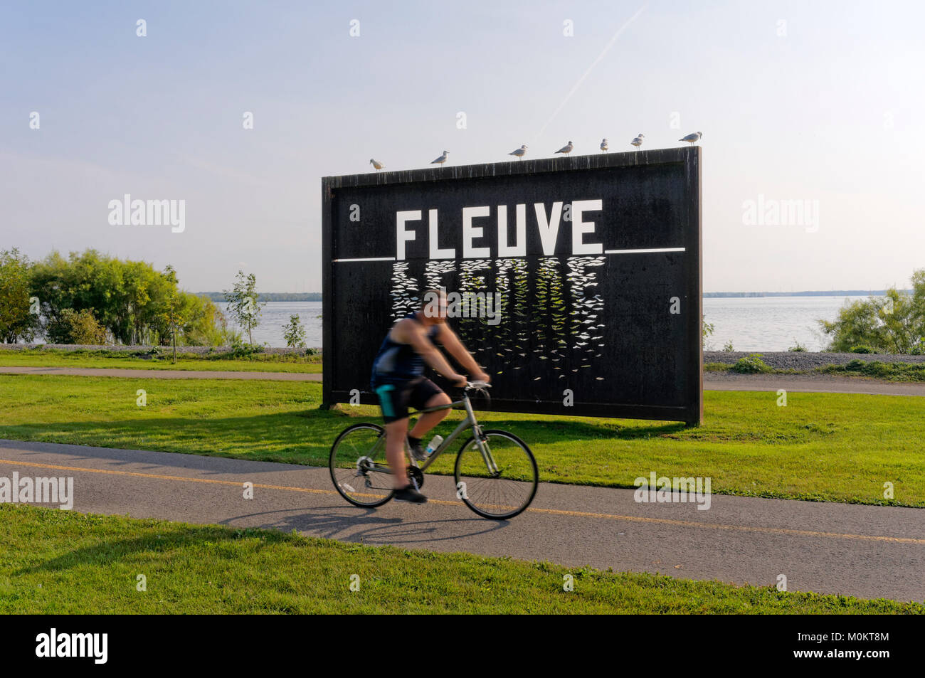 Uomo in sella ad una bicicletta passato grande pannello in metallo Installazione artistica con il fiume San Lorenzo in background, Lachine, Montreal, Quebec, Canada Foto Stock
