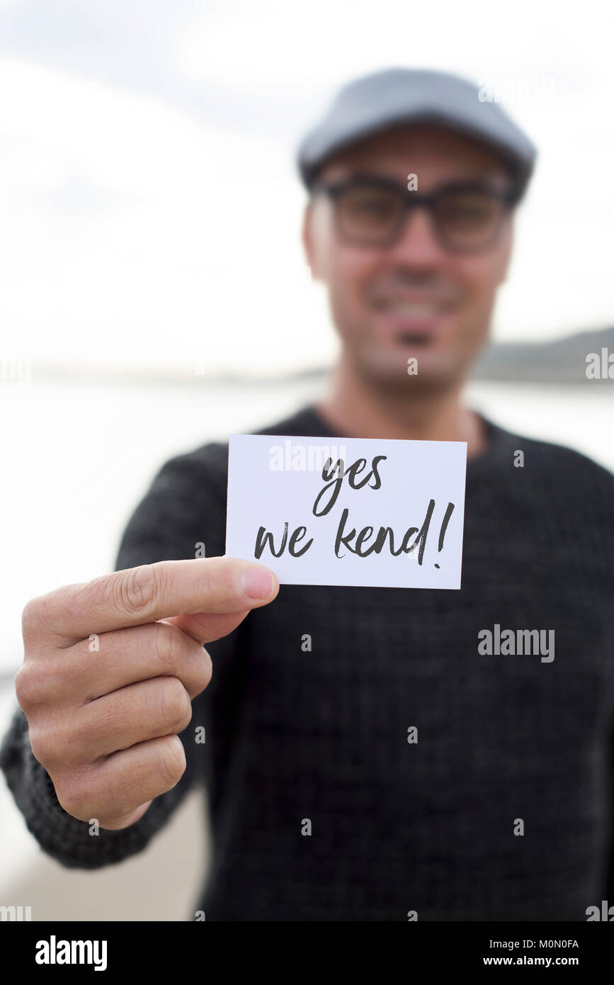 Primo piano di un giovane uomo caucasico indossando occhiali da vista e da un tappo piatto all'aperto mostra un cartello con scritto il testo yes we kend scritto in essa come yes we can Foto Stock