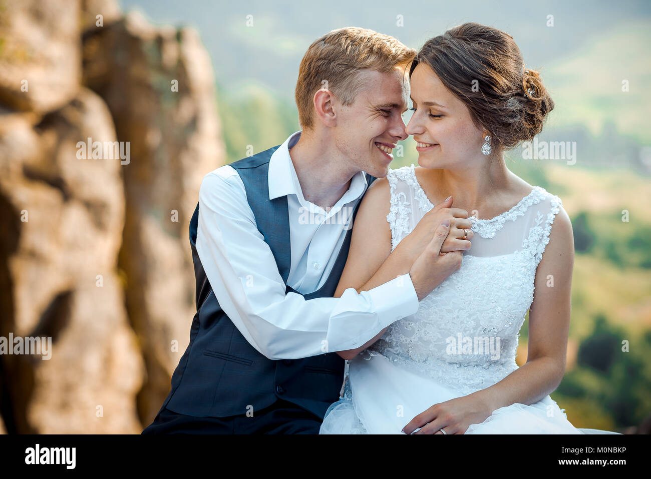 Emotiva ritratto di nozze. Giovane e bella sposa giovane è abbastanza sorridente, teneramente per mano e lo sfregamento dei nasi seduti sulle rocce. Foto Stock