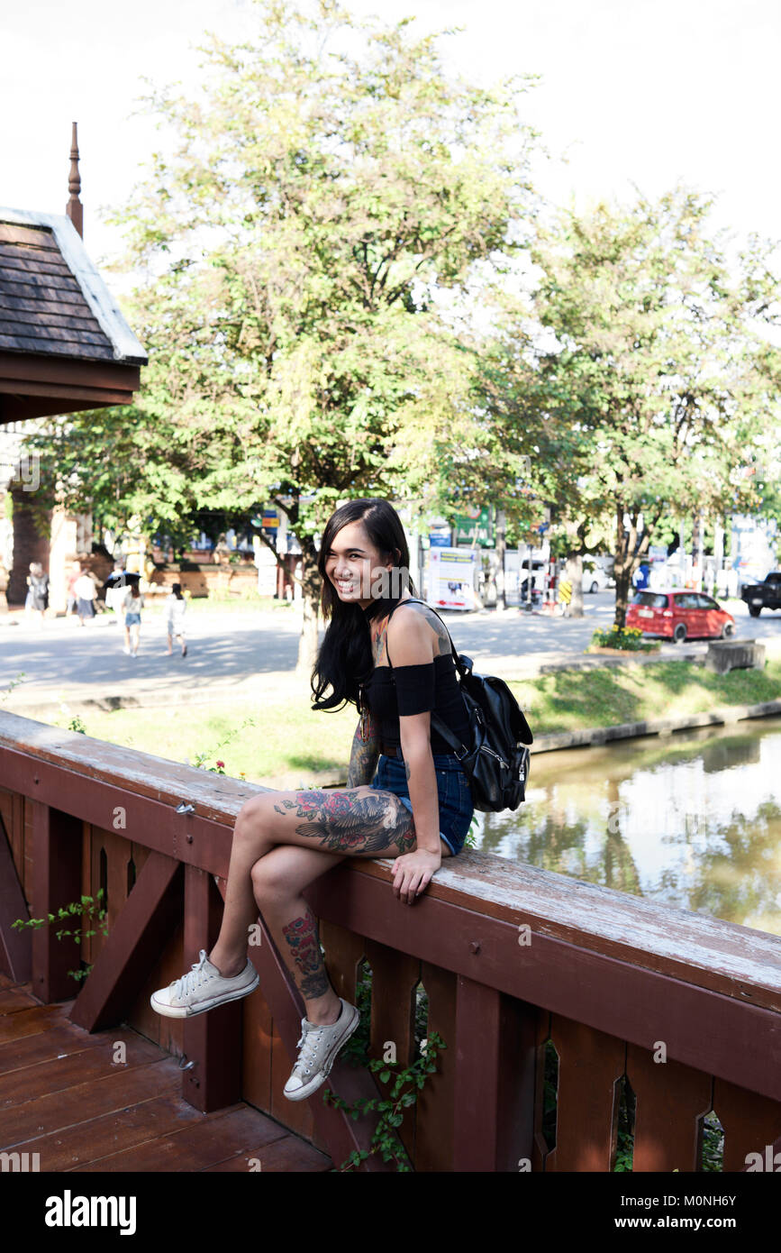 Tatuato brunnete asiatica ragazza sorridente alla telecamera mentre sitted su un ponte di legno in estate. Foto Stock