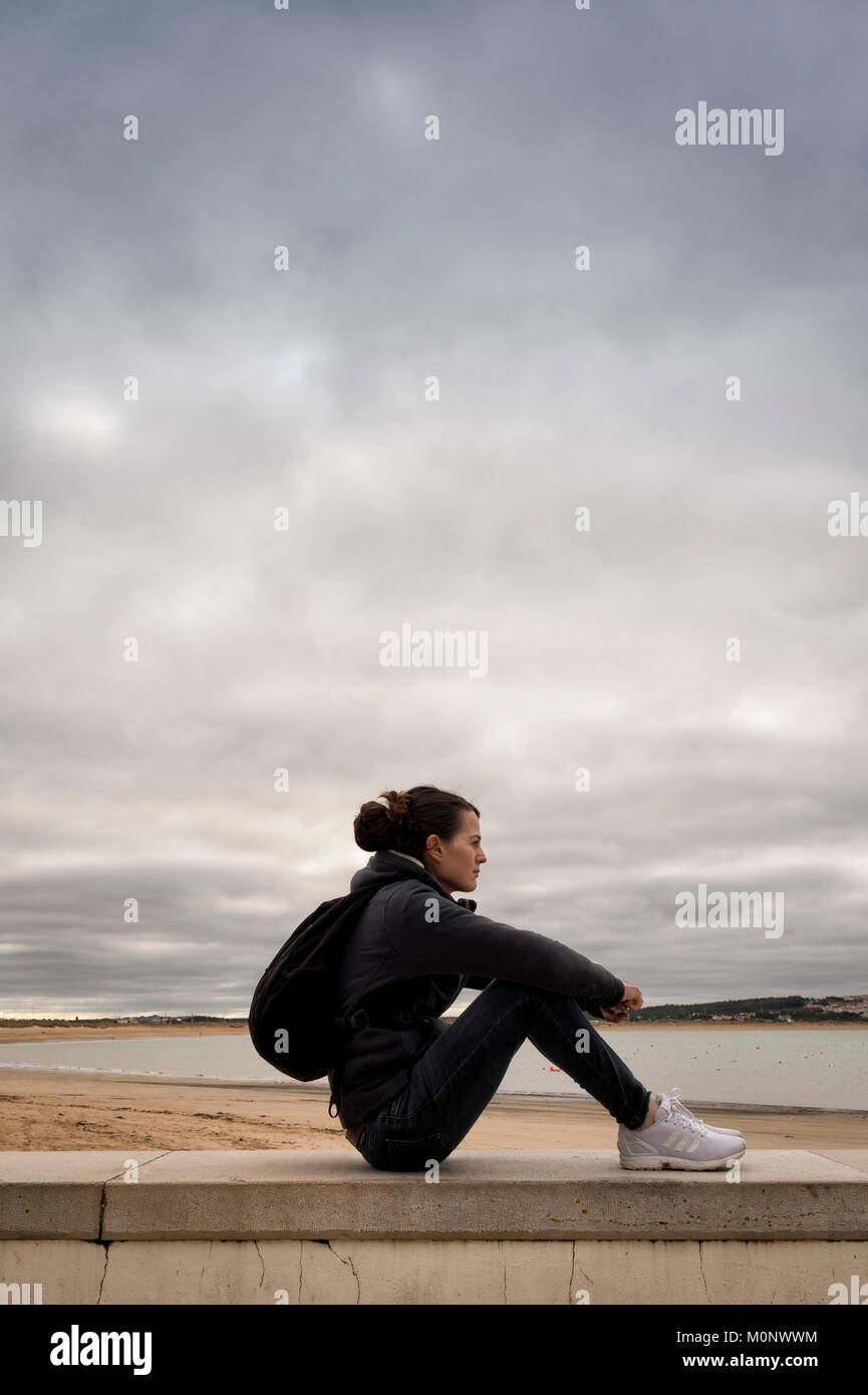 Donna seduta su un muro accanto al mare, indossando uno zaino di riposo. Foto Stock