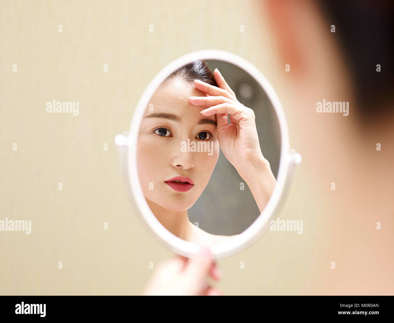 Bella giovane donna asiatica guardando a sé nel mirror, mano sulla fronte. Foto Stock