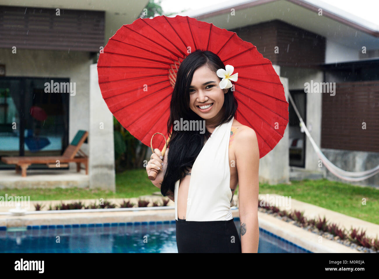 Pretty asian brunette millenaria tenendo un rosso ombrellone tradizionale accanto alla piscina di sorridere alla telecamera. Foto Stock