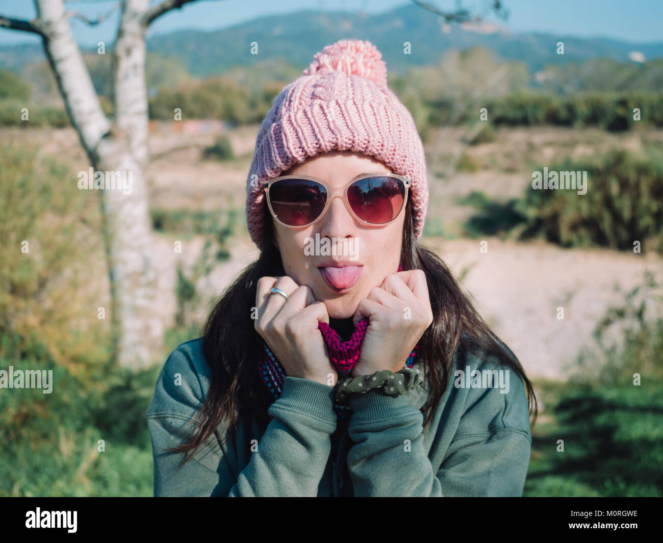 Donna felice con la lingua fuori all'aperto in inverno con un cappello di lana e occhiali da sole Foto Stock