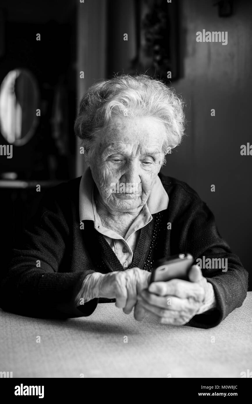 Vecchia donna prende sullo smartphone seduti a tavola in casa. In bianco e nero photo. Foto Stock