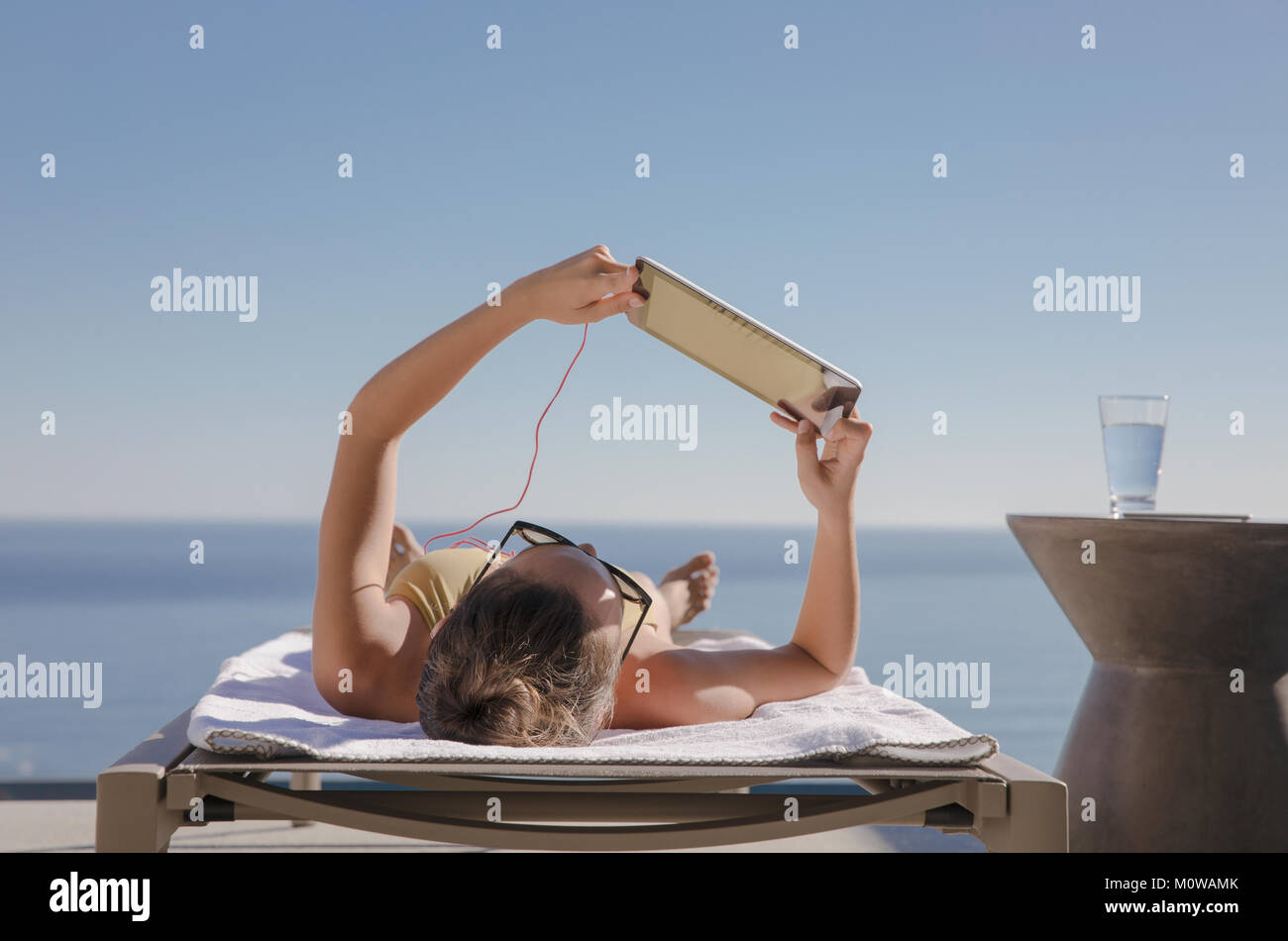 La donna a prendere il sole, con tavoletta digitale sulla sedia a sdraio sul patio soleggiato con vista oceano Foto Stock