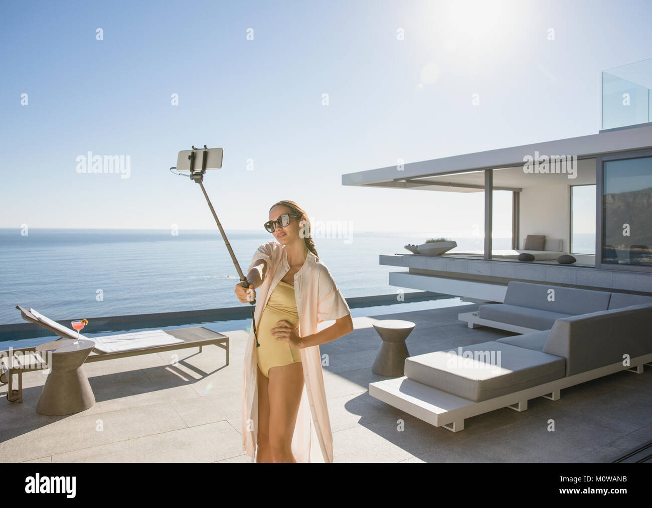 Donna in costume da bagno tenendo selfie con selfie stick sulla soleggiata, moderno luxury home vetrina patio esterno con vista oceano Foto Stock