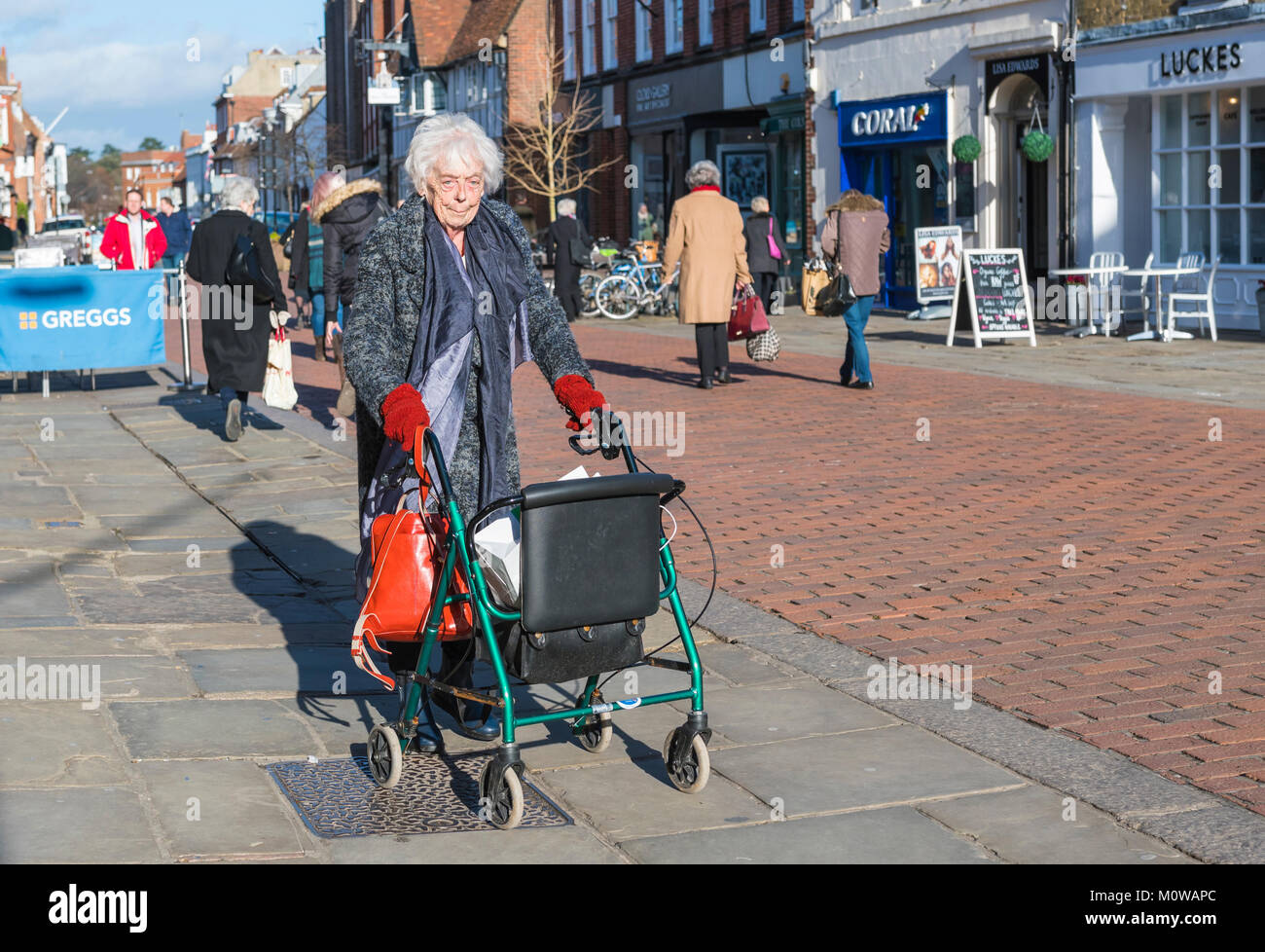Donna anziana con rollator su ruote (gommato zimmer frame aiuti a piedi) in un pedestrianise area dello shopping in Chichester, West Sussex, in Inghilterra, Regno Unito. Foto Stock