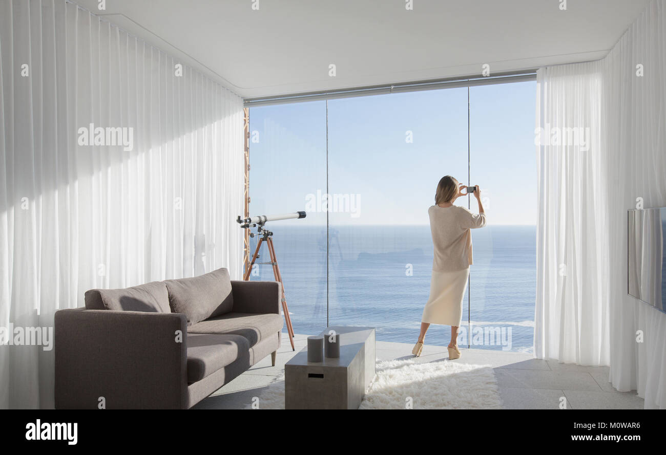 Donna con la fotocamera del telefono a fotografare soleggiata vista oceano dalla moderna, lusso home vetrina interno soggiorno Foto Stock