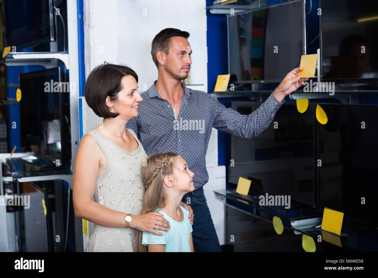 Coppia sorridente con piccola figlia scegliendo nuovo televisore in un negozio di elettrodomestici. Focus sulla donna Foto Stock
