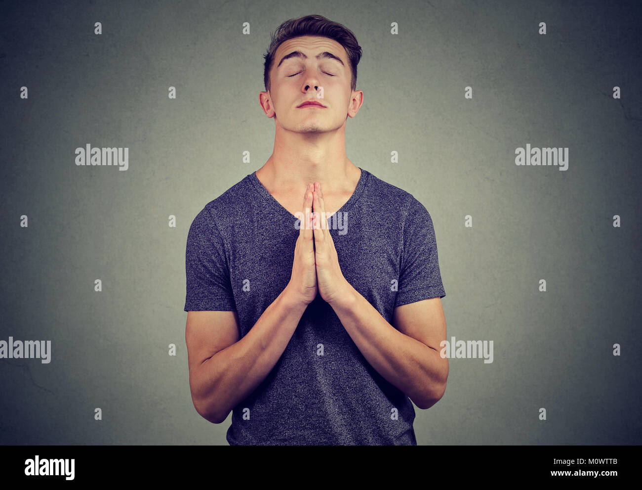 Giovane uomo informale tenendo le mani insieme a pregare nella fede mantenendo gli occhi chiusi. Foto Stock