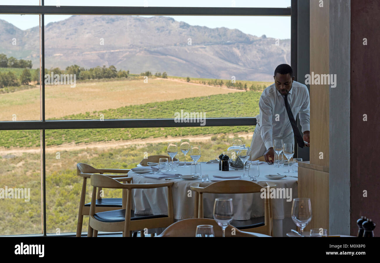 Western Cape regione Sud Africa. Circa 2017. Cameriere che stabilisce una tabella di ristorante che ha una vista panoramica di vigne e montagne Foto Stock