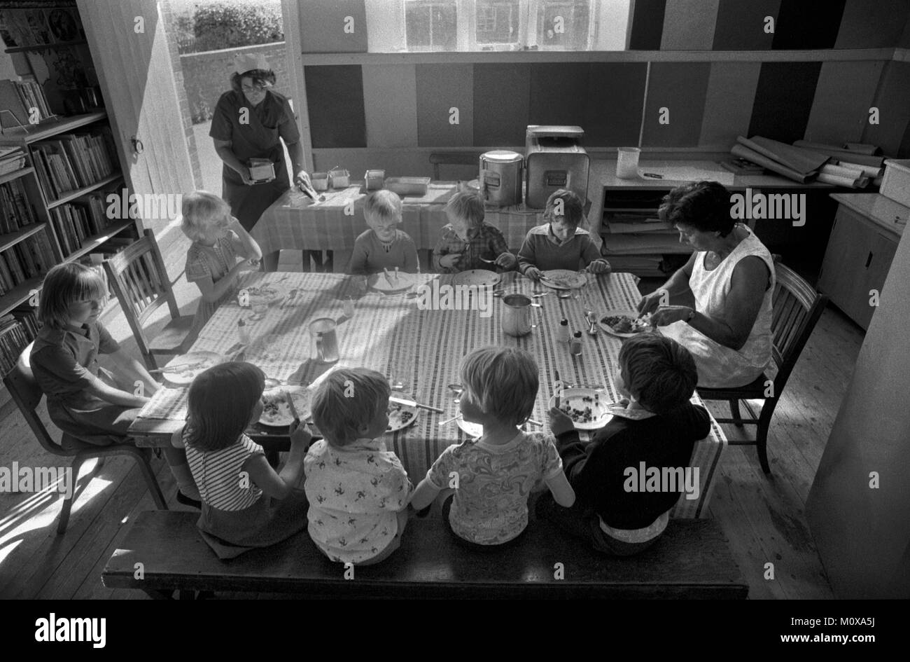 Village Scuola primaria 1970 Inghilterra. I bambini della scuola siedono insieme a un tavolo con un membro del personale e pranzano. Scuola cena signora in cappello bianco da tavolo lungo. Cheveley Cambridgeshire 1978 70s UK HOMER SYKES Foto Stock