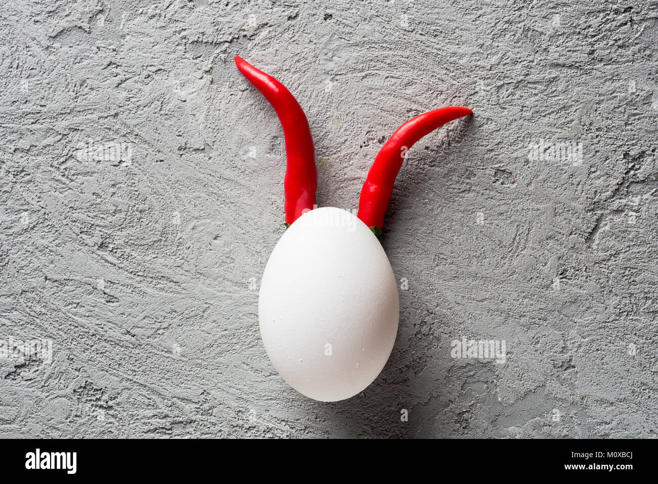 Il guscio delle uova e il red hot chili pepper imitazione faccia del diavolo clown su pietra grigia background paper. Concetto di alimentare il minimo Halloween Foto Stock