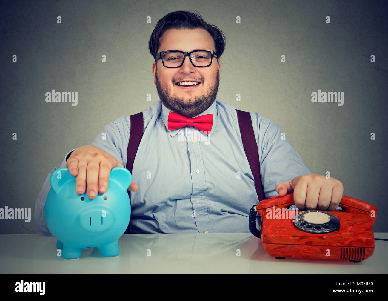 Giovane uomo sorridente in posa con il salvadanaio e rosso vecchio telefono lavora in banca. Foto Stock