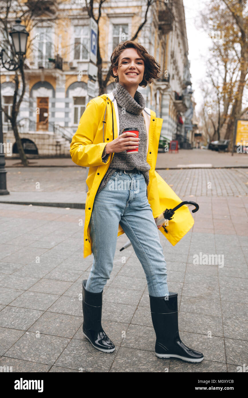 Foto di allegro piuttosto giovane donna vestita in stivali di gomma e  impermeabile giallo passeggiate all'aperto a bere caffè. Guardando la  fotocamera Foto stock - Alamy