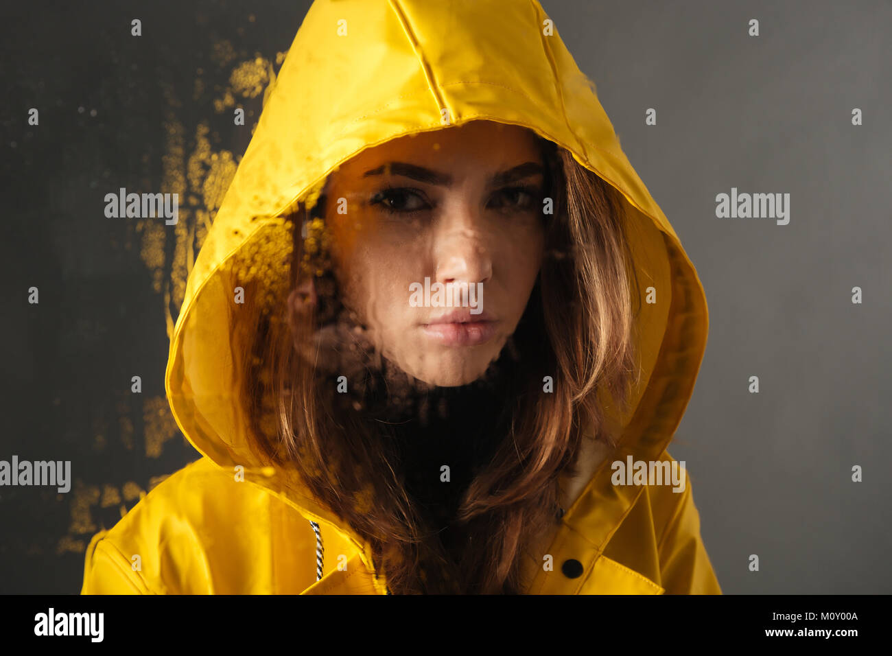 Close up di un sconvolto ragazza vestita in impermeabile con un cappuccio sulla sua testa in piedi dietro un umido con finestra pioggia isolate su uno sfondo grigio Foto Stock