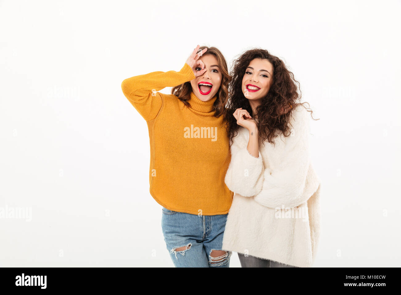 Due ragazze gioiosa in maglioni per divertirsi insieme e guardando la telecamera su sfondo bianco Foto Stock