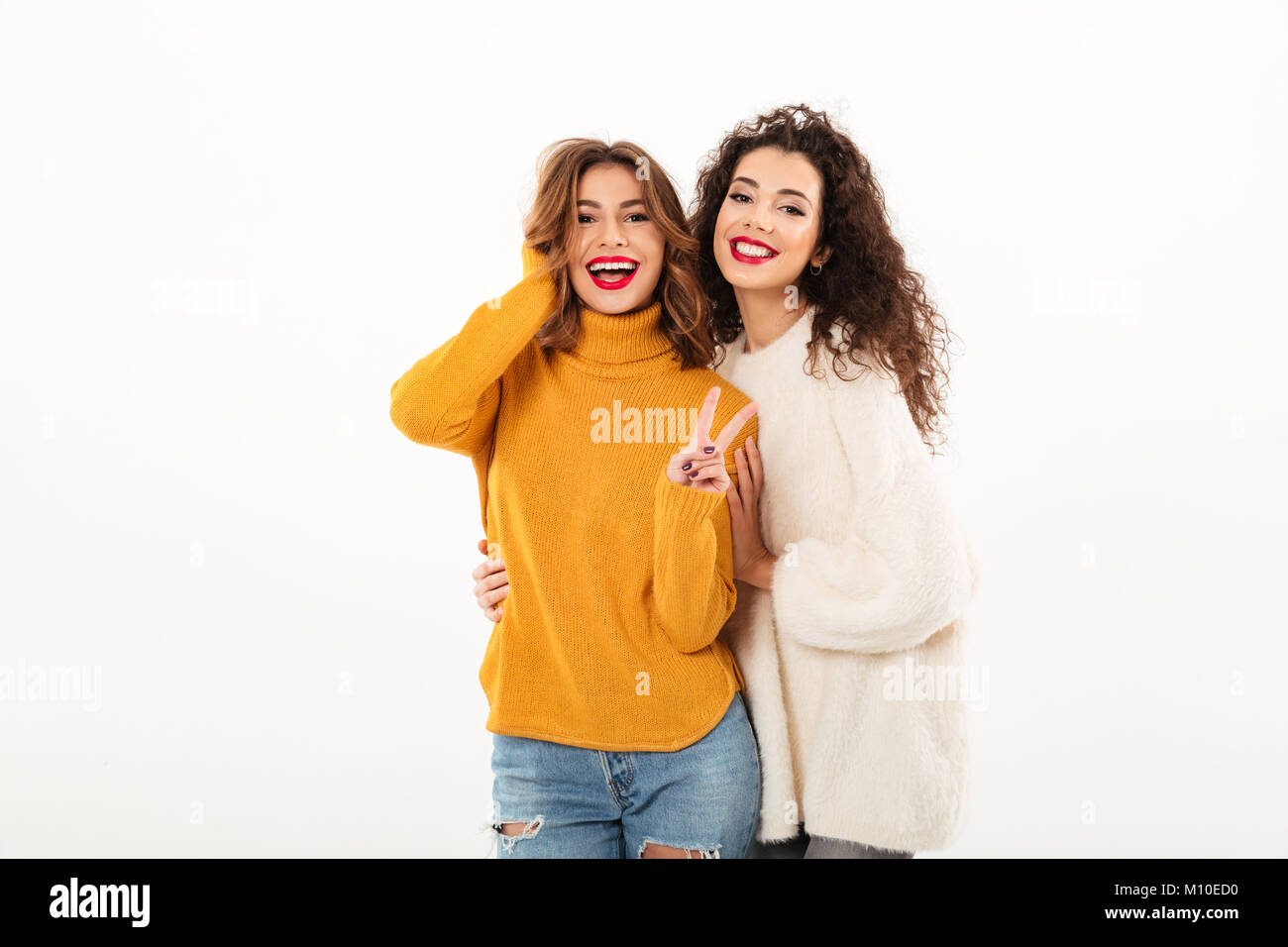 Due ragazze sorridenti in maglioni in posa insieme e guardando la telecamera mentre una donna che mostra il gesto di pace su sfondo bianco Foto Stock