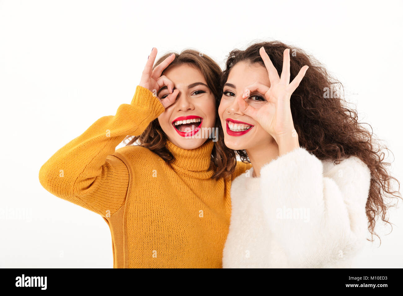 Immagine di due ragazze felici in maglioni divertendosi e mostrando i gesti ok mentre guardando la telecamera su sfondo bianco Foto Stock