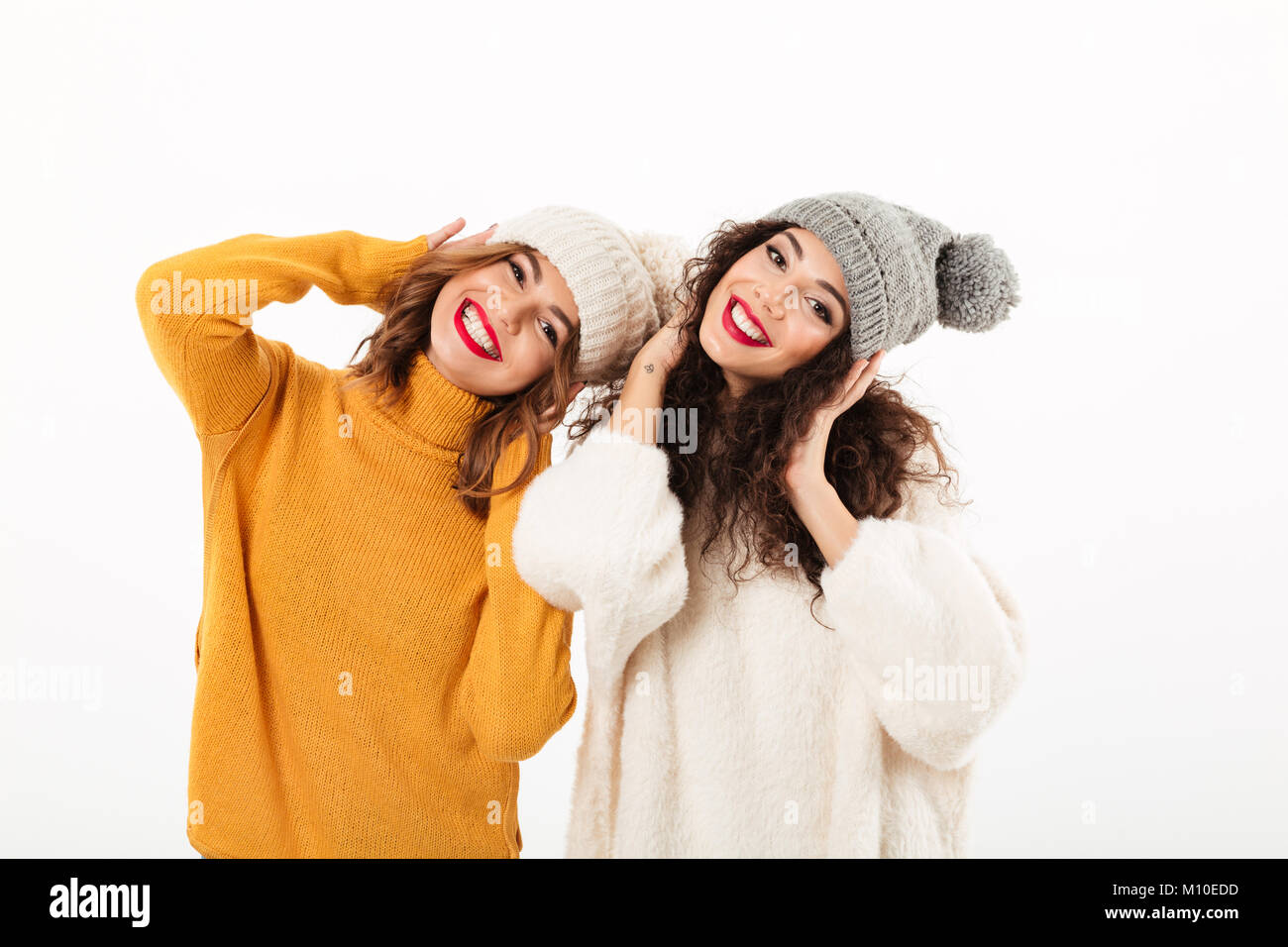Due ragazze felici in maglioni e cappelli per divertirsi insieme mentre guardando la telecamera su sfondo bianco Foto Stock