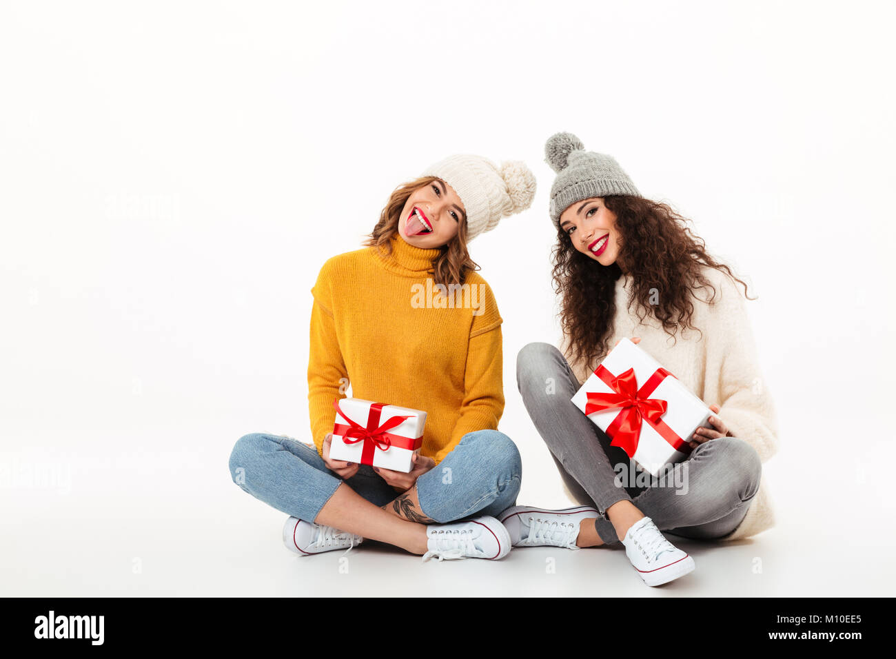 Due ragazze giocoso in maglioni e cappelli seduta con regali sul pavimento insieme mentre guardando la telecamera su sfondo bianco Foto Stock