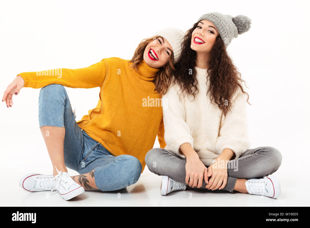 Due ragazze felici in maglioni e cappelli di seduta sul pavimento insieme e guardando la telecamera su sfondo bianco Foto Stock