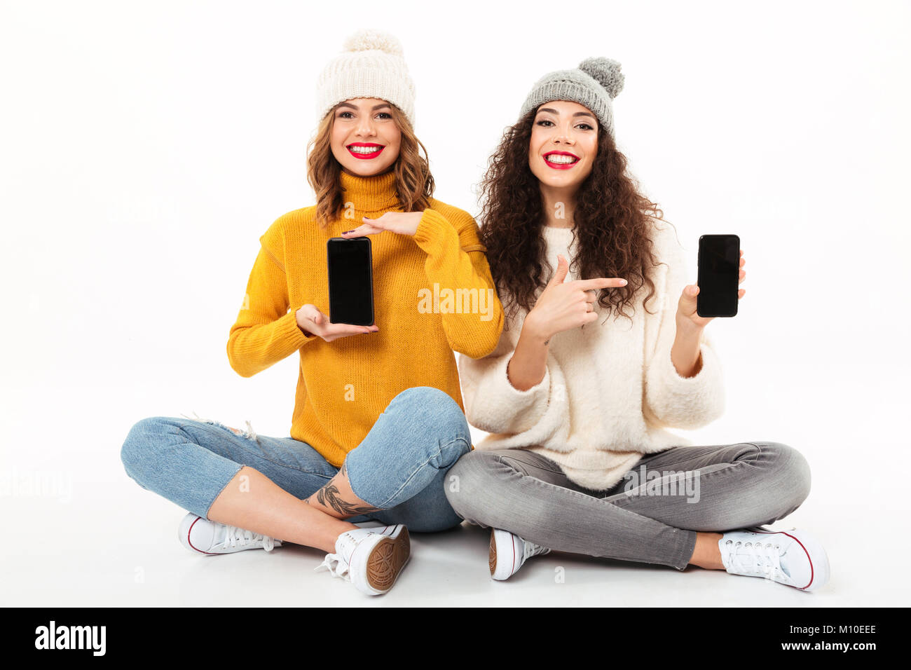 Due ragazze felici in maglioni e cappelli di seduta sul pavimento insieme mentre mostra gli smartphone sbozzati schermate e guardando la telecamera su bianco backg Foto Stock