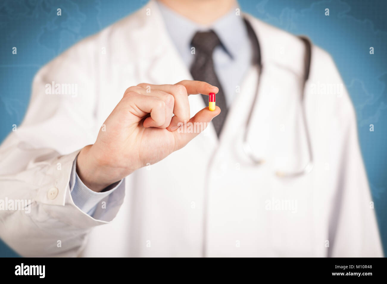 Maschio di un medico in camice bianco davanti a una parete blu, con uno stetoscopio su uno spallamento tenendo una pillola tra le sue dita Foto Stock