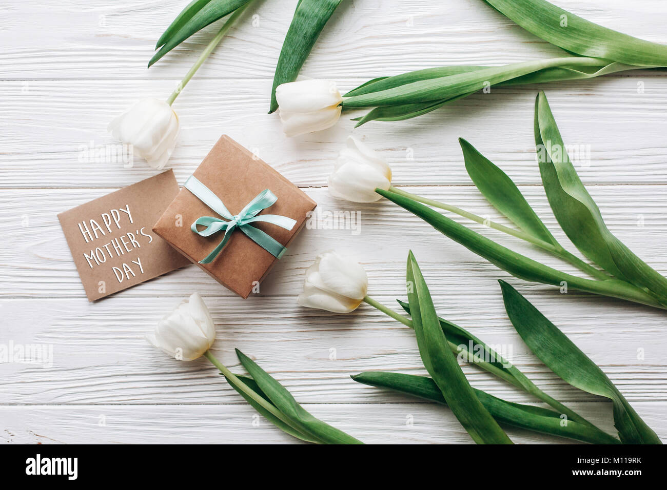 Felice Festa della mamma segno di testo su elegante imbarcazione presente casella e biglietto di auguri e tulipani su legno bianco sfondo rustico. laici piatto con fiori e spa Foto Stock