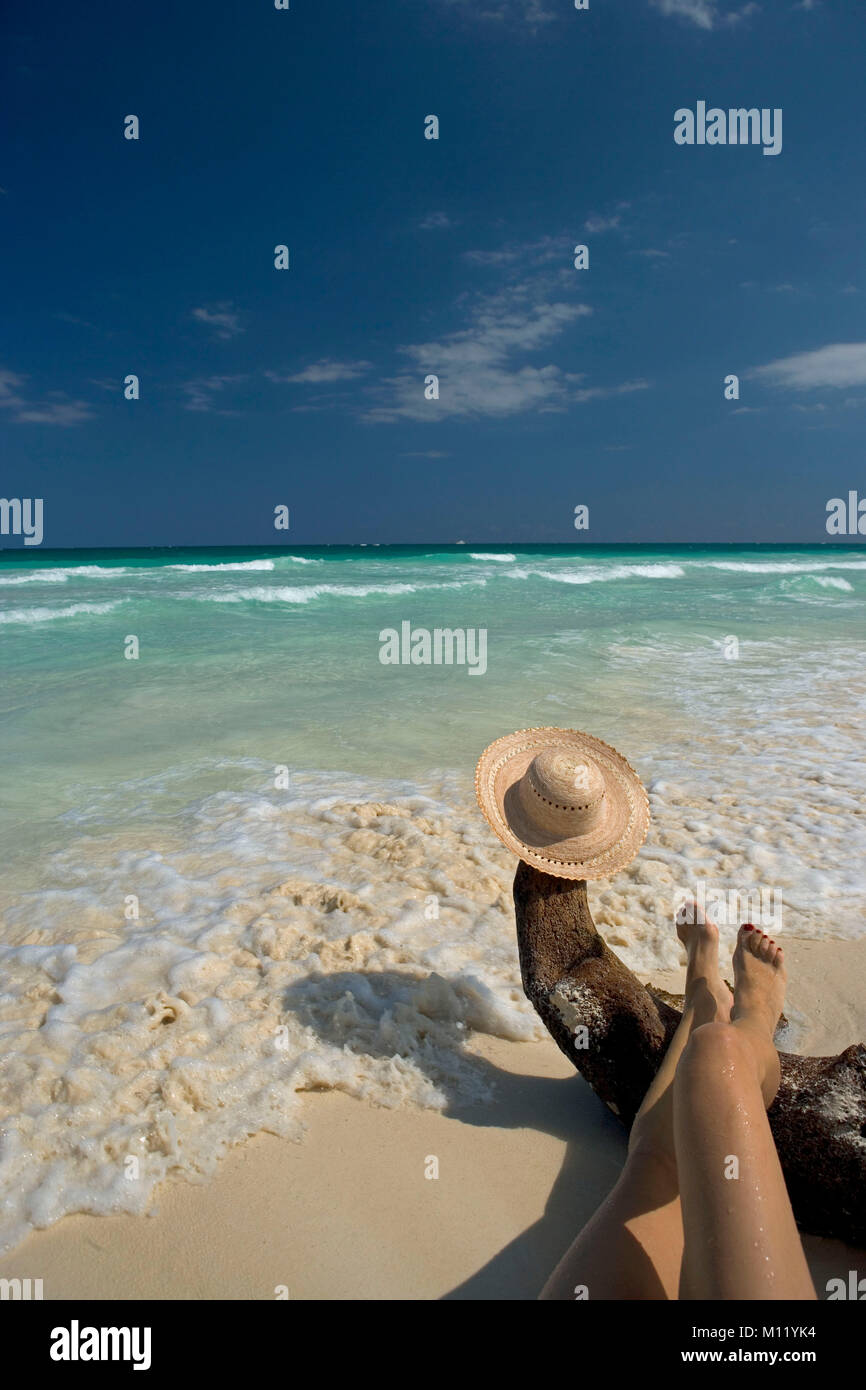 Messico. La penisola dello Yucatan. Tulum. Cappello per il sole e il turista femminile gambe in appoggio sul driftwood sulla spiaggia. Foto Stock