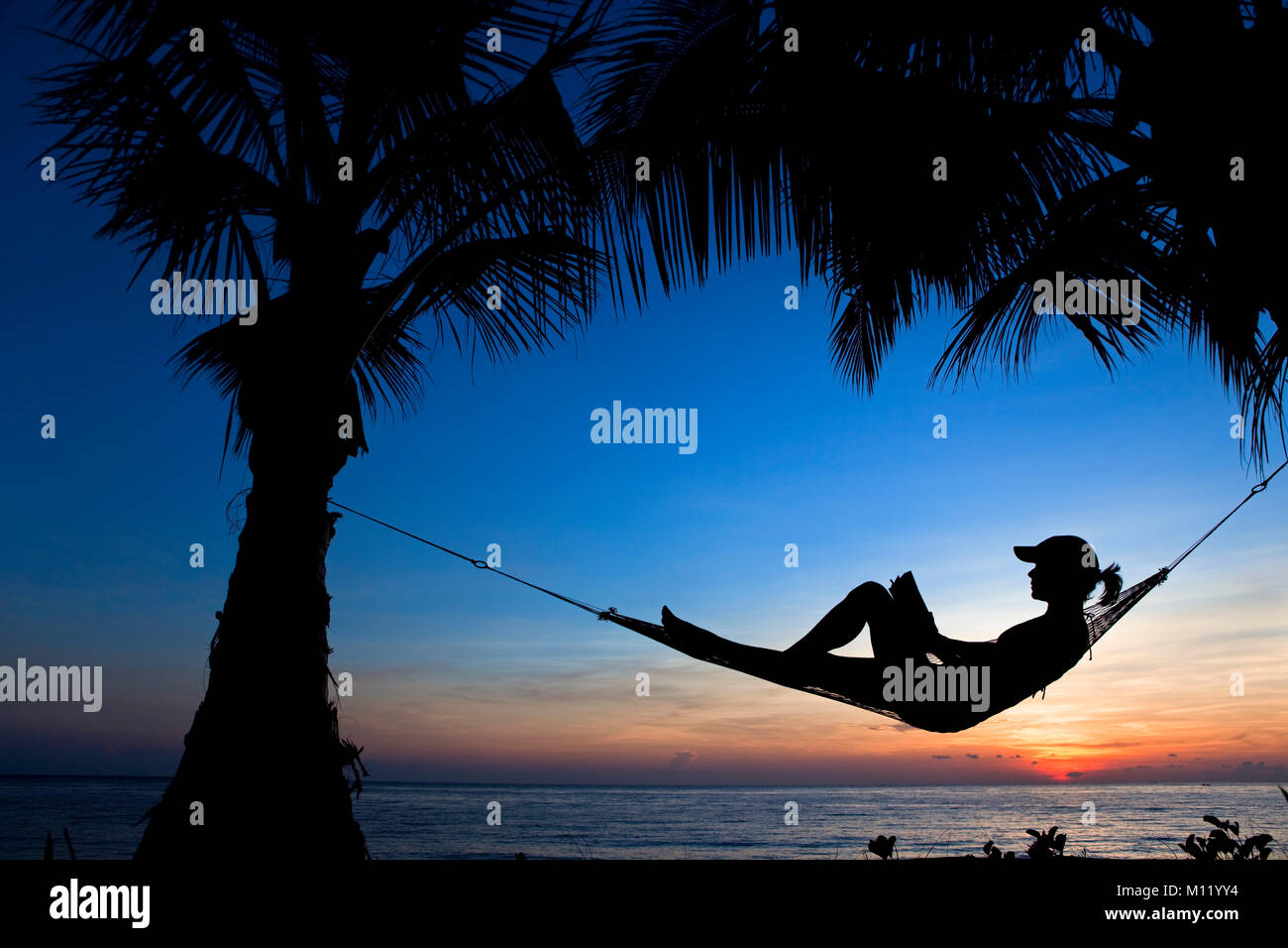Indonesia, isola di Bali, vicino al villaggio di Tejakula, Gaia Oasis Resort. Donna libro lettura in amaca al tramonto. Foto Stock