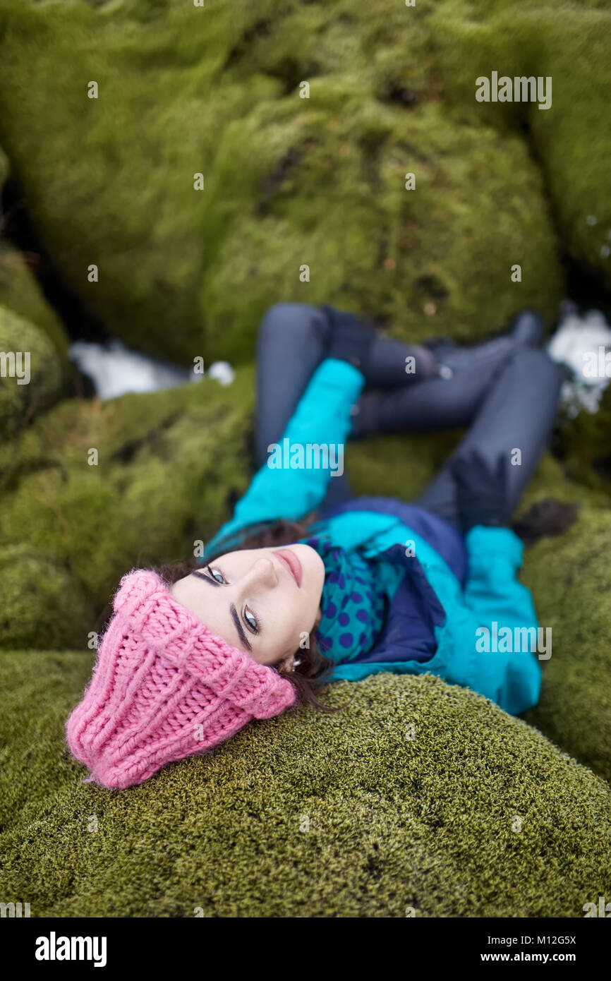 Deliziosa ragazza giace sul verde muschio rock in Islanda. Indossa una  giacca blu con pantaloni neri, guanti e un cappello rosa. Donna guarda  verso la fotocamera w Foto stock - Alamy