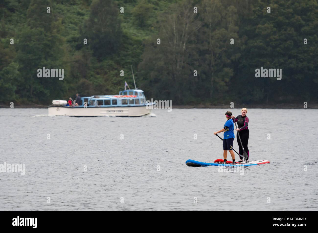 2 Femmine stand su paddleboards sul Coniston Water mentre la gente sul lancio crociera in barca Gita in barca a vela sono al di là - Lake District, Cumbria, Inghilterra, Regno Unito. Foto Stock