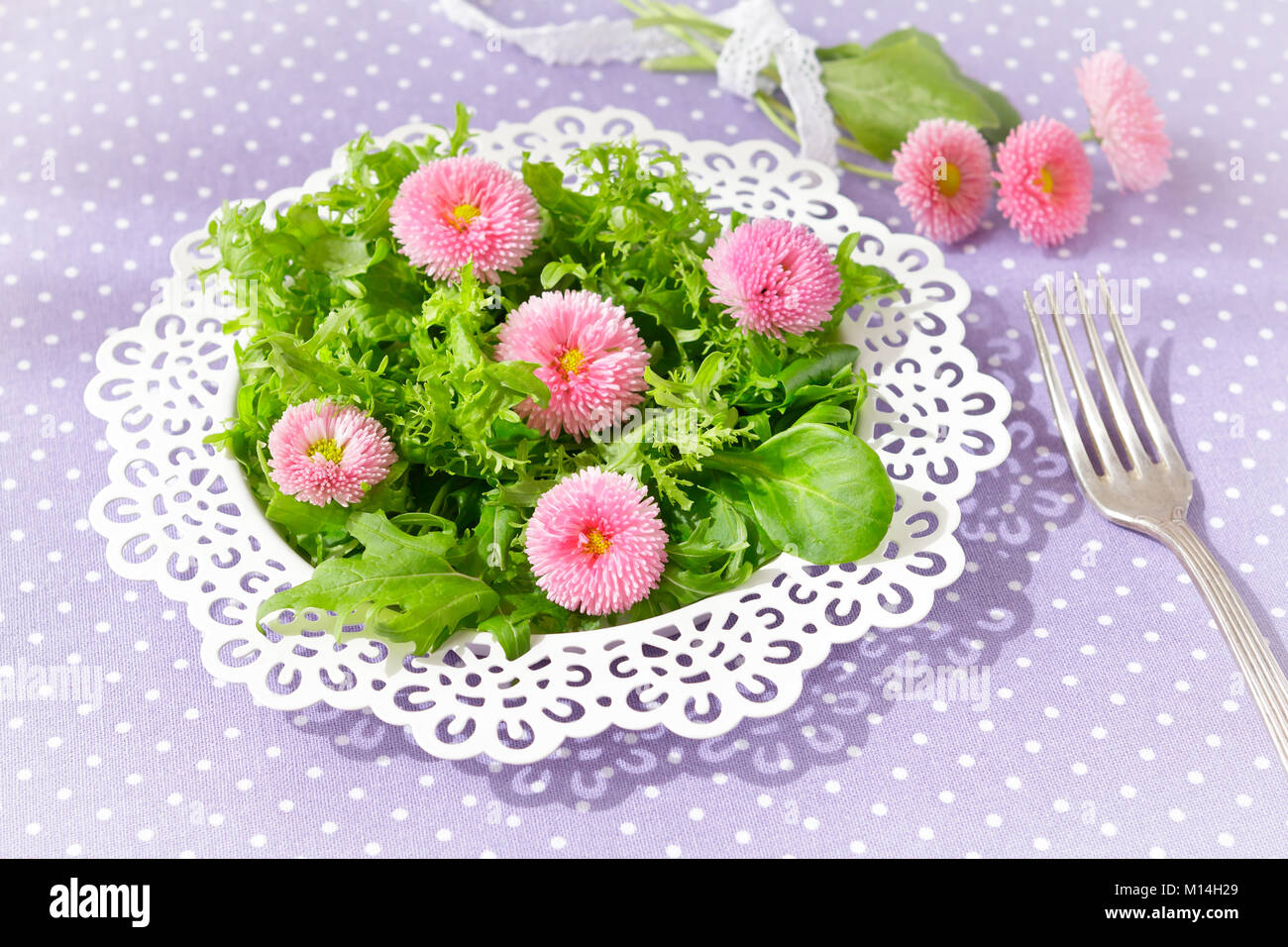 Piastra bianca con una insalata verde mista le foglie di lattuga e commestibili fiori a margherita, insieme con una forcella di vintage su un nostalgico sfondo lilla Foto Stock