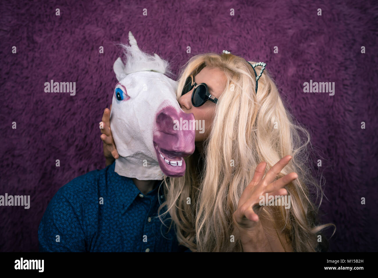 Freaky giovane donna kissing felice unicorn sullo sfondo viola. Bionda espressiva con strano ragazzo. Insolito ci si siede insieme Foto Stock