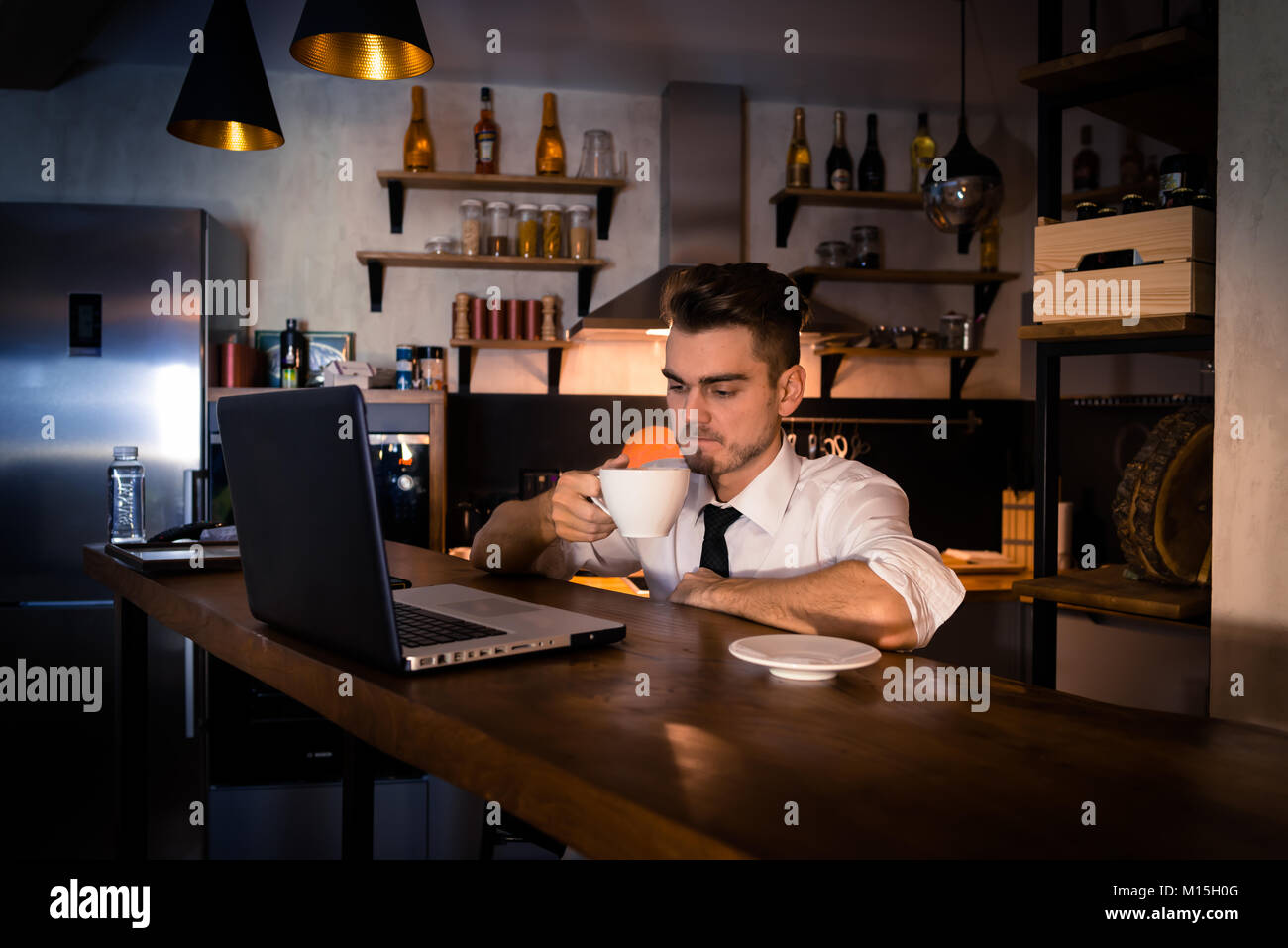 Giovane uomo si siede in cucina al bancone del bar lavora in laptop e beve un caffè. Atmosfera di casa, lavoro di sera dopo una dura giornata Foto Stock