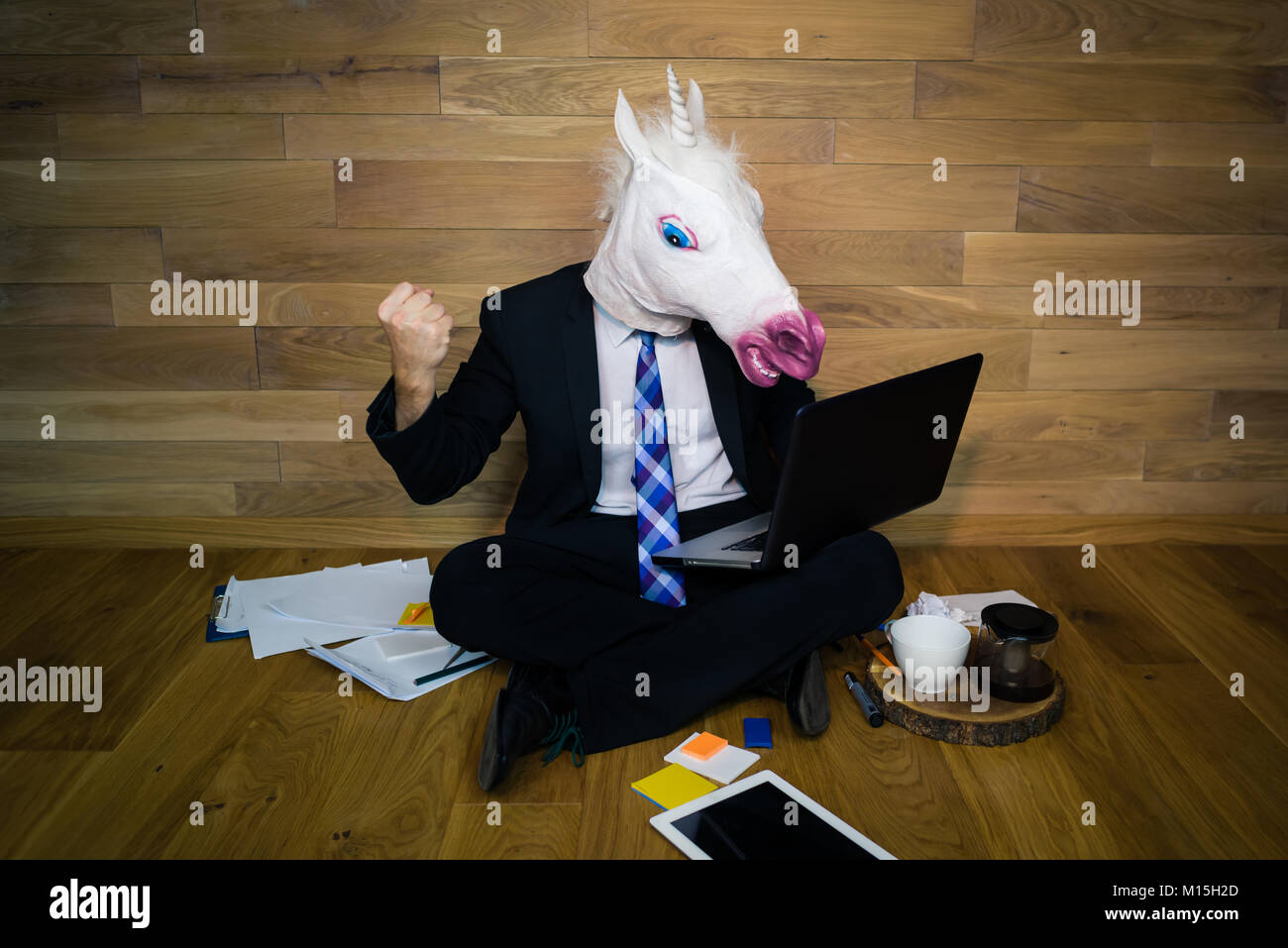 Giovane uomo nel divertente maschera di cavallo si siede sul pavimento con la tazza di caffè e lavora con il computer portatile. Arrabbiato e insoddisfatto unicorn in una tuta mostra pugno Foto Stock