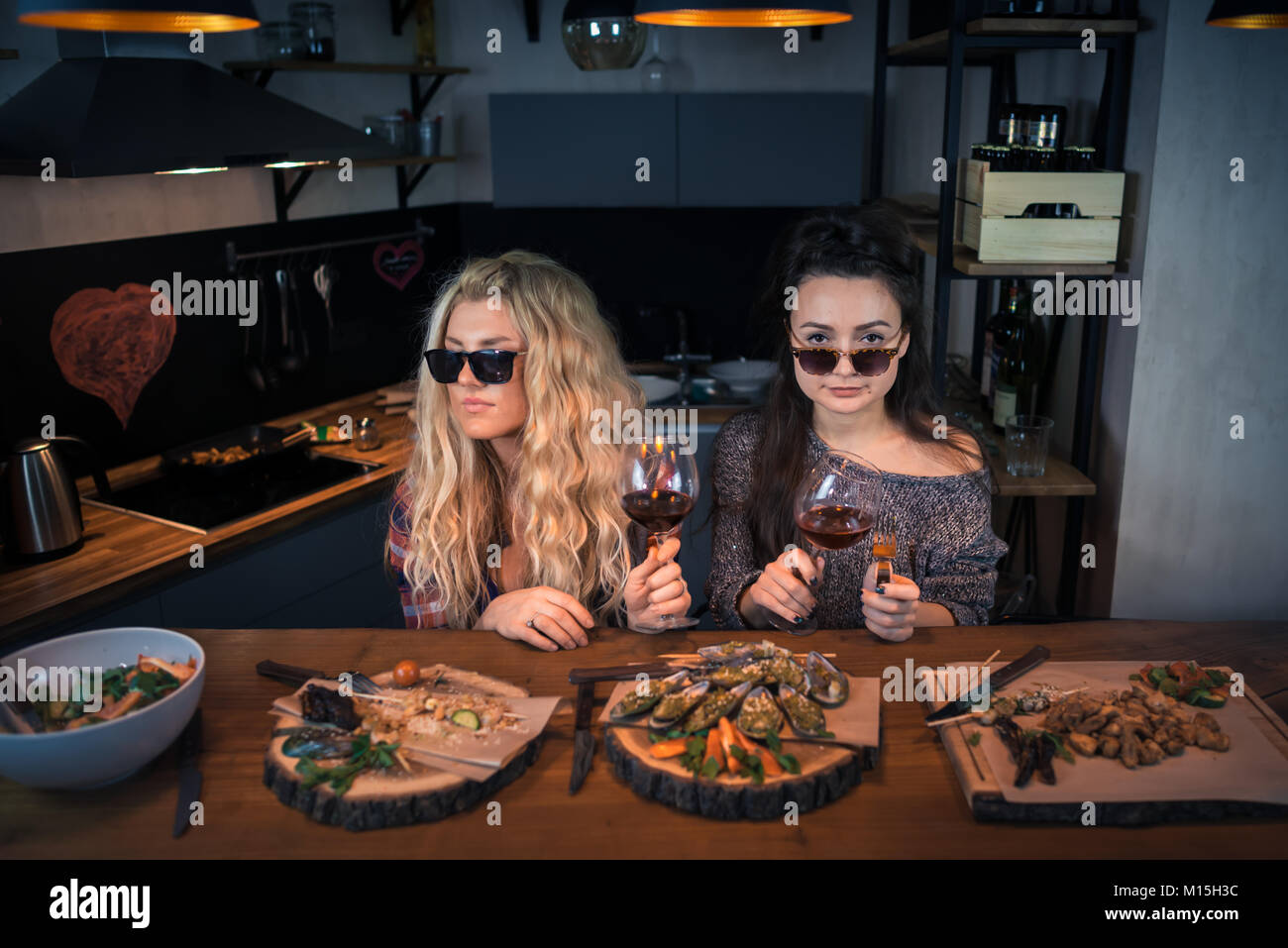 Due giovani donne detiene i bicchieri di vino in casa moderna cucina. La gente si siede a tavola con gli alimenti. Fidanzate godendo di sera insieme Foto Stock