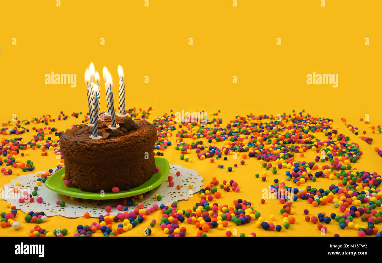 Cioccolato torta di compleanno con illuminato blu e candele bianche, su una piccola piastra verde circondato da sfere di caramella disseminata su sfondo giallo Foto Stock