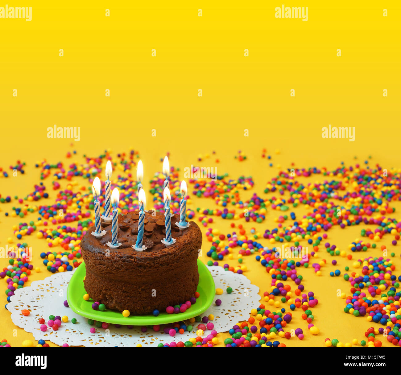 Cioccolato torta di compleanno con illuminato blu e candele bianche, su una piccola piastra verde circondato da sfere di caramella disseminata su sfondo giallo Foto Stock