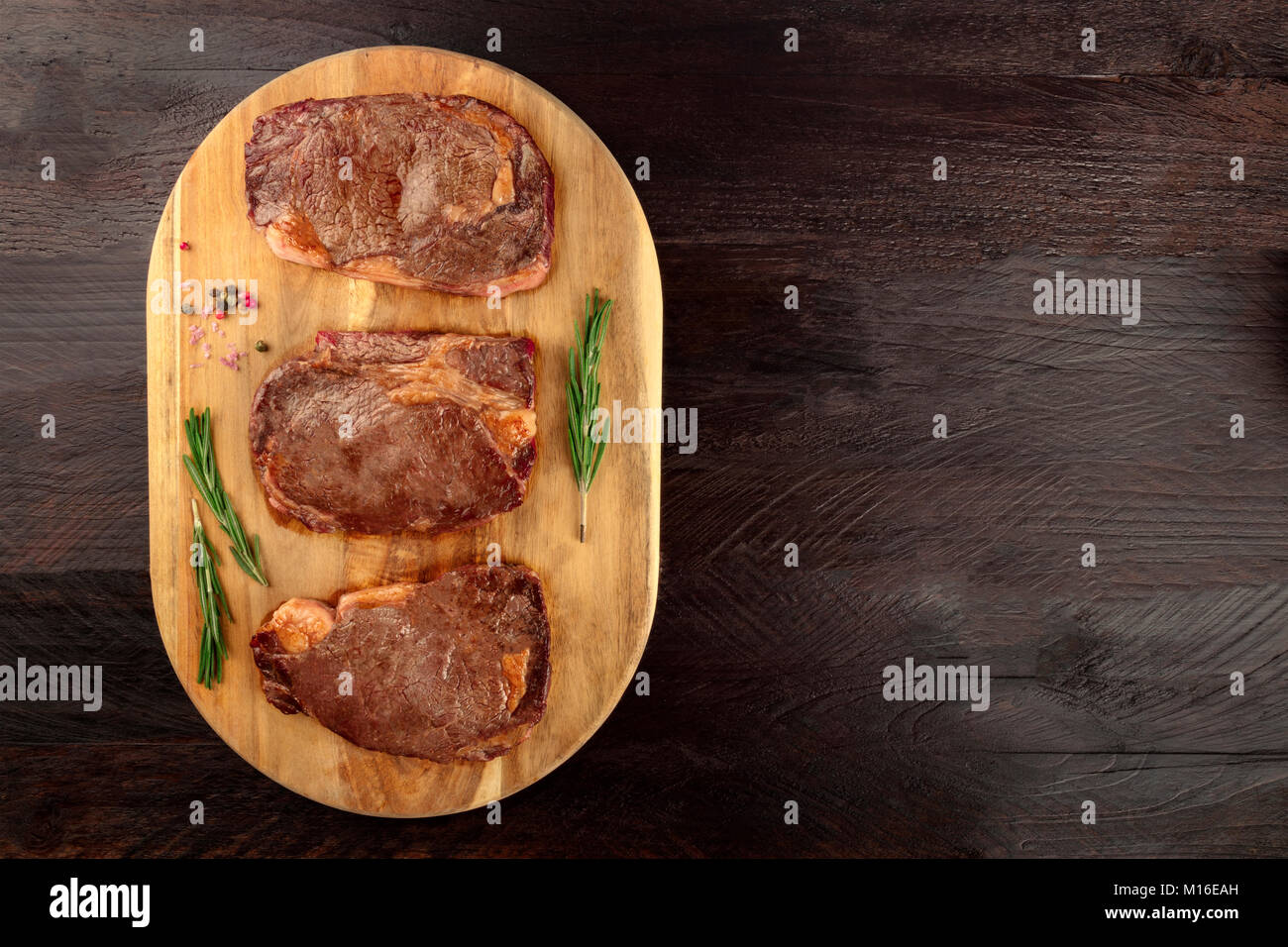 Una foto aerea di tre fette di carne cotta, filetti di manzo, ripresa dall'alto sul buio rustica con rametti di rosmarino, sale, pepe e un p Foto Stock