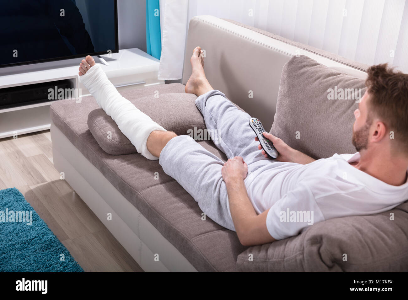Giovane uomo Con gamba rotta sdraiato sul divano Azienda Remote vicino al televisore Foto Stock