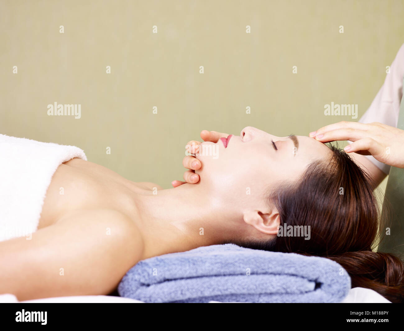 Vista laterale di una giovane e bella donna asiatica ricevere il massaggio del viso in spa salone a occhi chiusi. Foto Stock